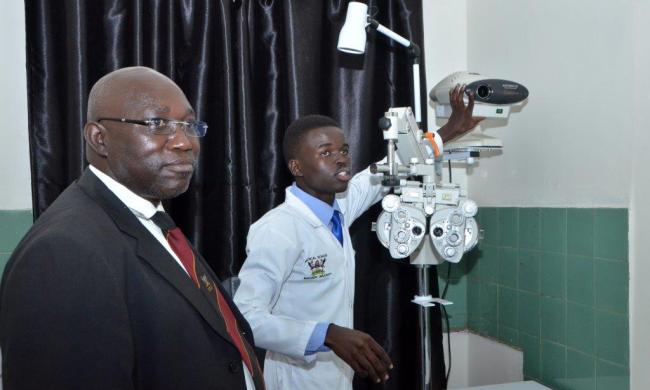 A student (in white coat) demonstrates to the College of Health Sciences (CHS) Deputy Principal-Dr. Isaac Okullo Visual acuity testing at the Optometry & Visual Science Centre, Makerere University Hospital, Kampala Uganda.