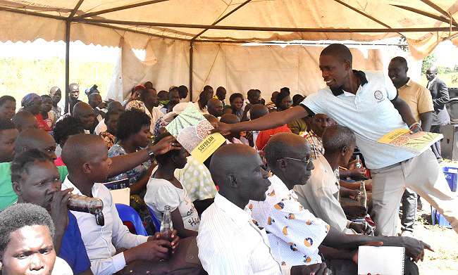 The Project Officer PASTTA Project Mr. Arnold Mbowa hands a copy of the Makerere University Soybean Production Guidebook to a farmer as one of the co-authors Mrs. Tonny Obua (2nd R) witnesses during the Field Day on 5th January 2019 at the Ngetta ZARDI, Lira District, Northern Uganda.