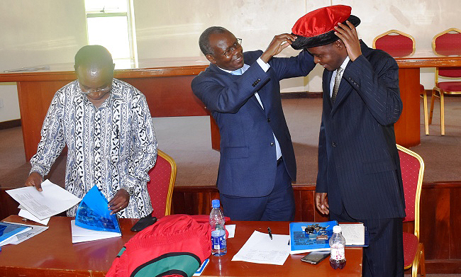 Principal CAES-Prof. Bernard Bashaasha (C) puts the Graduate Cap upon Dean-elect Dr. Abel Atukwase as outgoing Dean-Prof. John Muyonga (Left) prepares some handover material during the ceremony on 17th January 2019, Conference Hall, SFTNB, CAES, Makerere University, Kampala Uganda