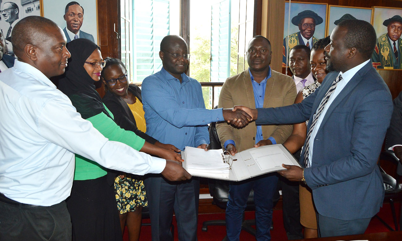 Ag. University Secretary-Mr. Yusuf Kiranda (Right) receives documents from KAPS’ Mr. Lawrence Madialo (4th L) as R-L: Eng. Christina Kakeeto, Mr. Evarist Bainomugisha, Mr. Samuel Mugabi, Ms. Nansubuga Jacqueline Anne, Ms. Fatuma Mohamed Ahmed and Mr. Kuria M. Njenga witness on 8th January 2020, Makerere University, Kampala Uganda.