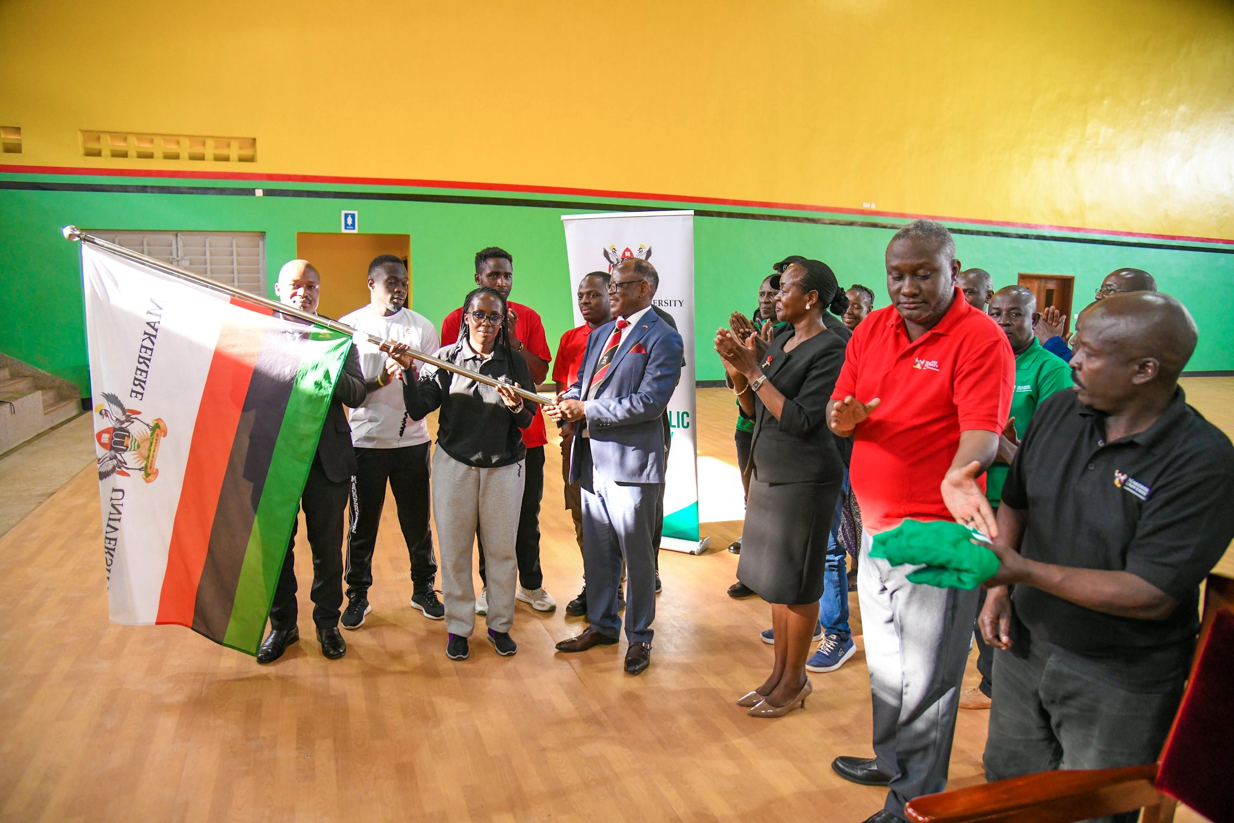 Prof. Barnabas Nawangwe hands the Makerere flag over to Team Captain-Namutebi Rhonah Majoline as Dr. Winifred Kabumbuli (3rd Right), Mr. Brian Miiro (2nd Right) and Left to Right: Dr. Rodney Rugyema, Phillip Nuwagaba, Katsirabo Donic, Akampa Bob James and others applaud on 17th December 2025 in the Mak Indoor Sports Arena. Prof. Barnabas Nawangwe officially flags off a contingent of 220 students, 13 coaches and 3 tutors from the Makerere University Games Union on 17th December 2025 at the Indoor Sports Arena to the 20th Edition of the Association of Uganda University Sports (AUUS) Games, 18th-23rd December 2025, Uganda Christian University (UCU) Mukono, Uganda, East Africa.