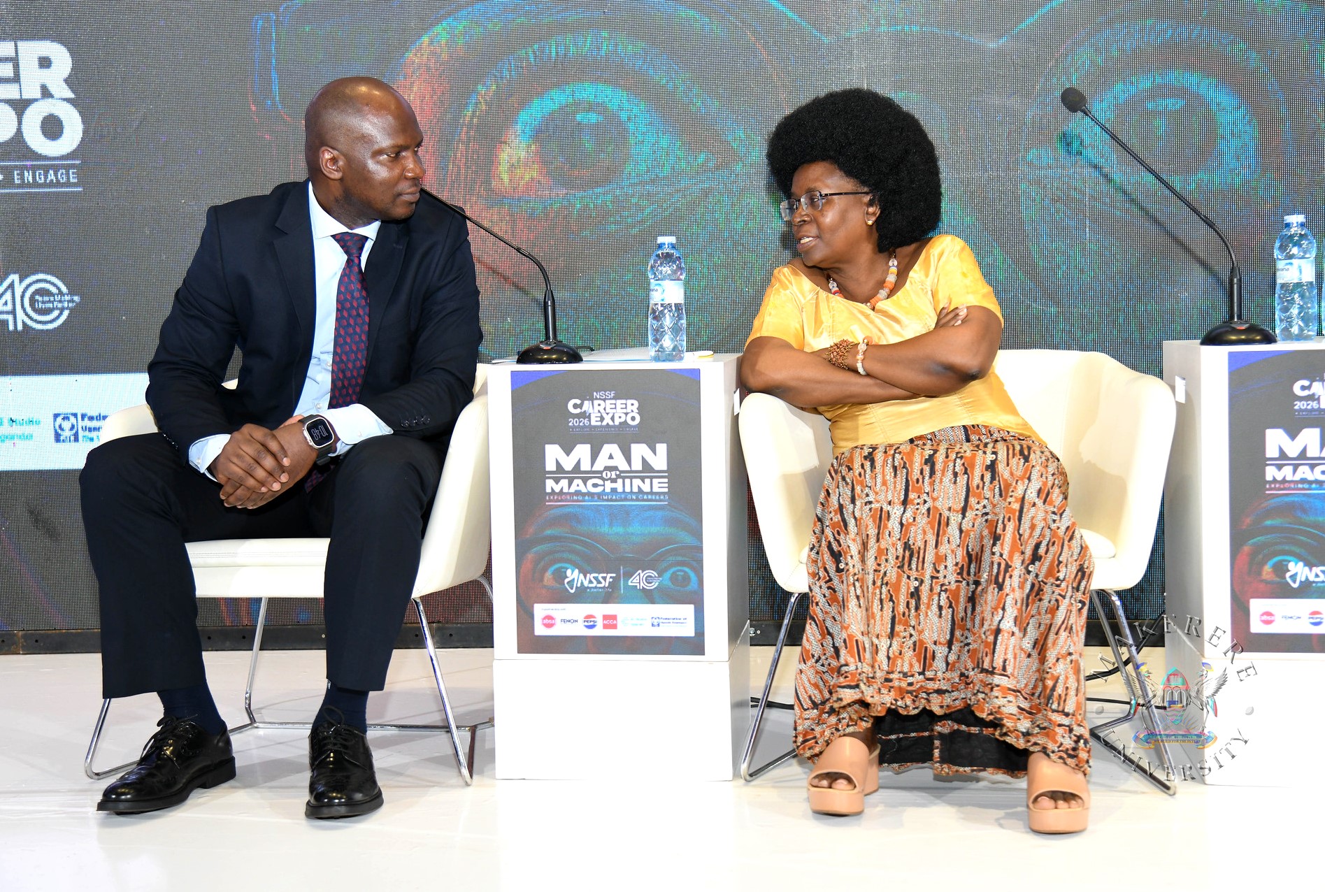 Hon. Dr. Monica Musenero (R) and NSSF's Gerald Paul Kasato (L) confer. Three-day Careers Fair 2026, bringing together students, industry leaders, and policymakers to discuss the future of work in the era of Artificial Intelligence (AI), Day 1 held under the theme “Machine vs. Man” and sponsored by the National Social Security Fund (NSSF), 11th March 2026, Yusuf Lule Central Teaching Facility Auditorium, Makerere University, Kampala Uganda, East Africa.