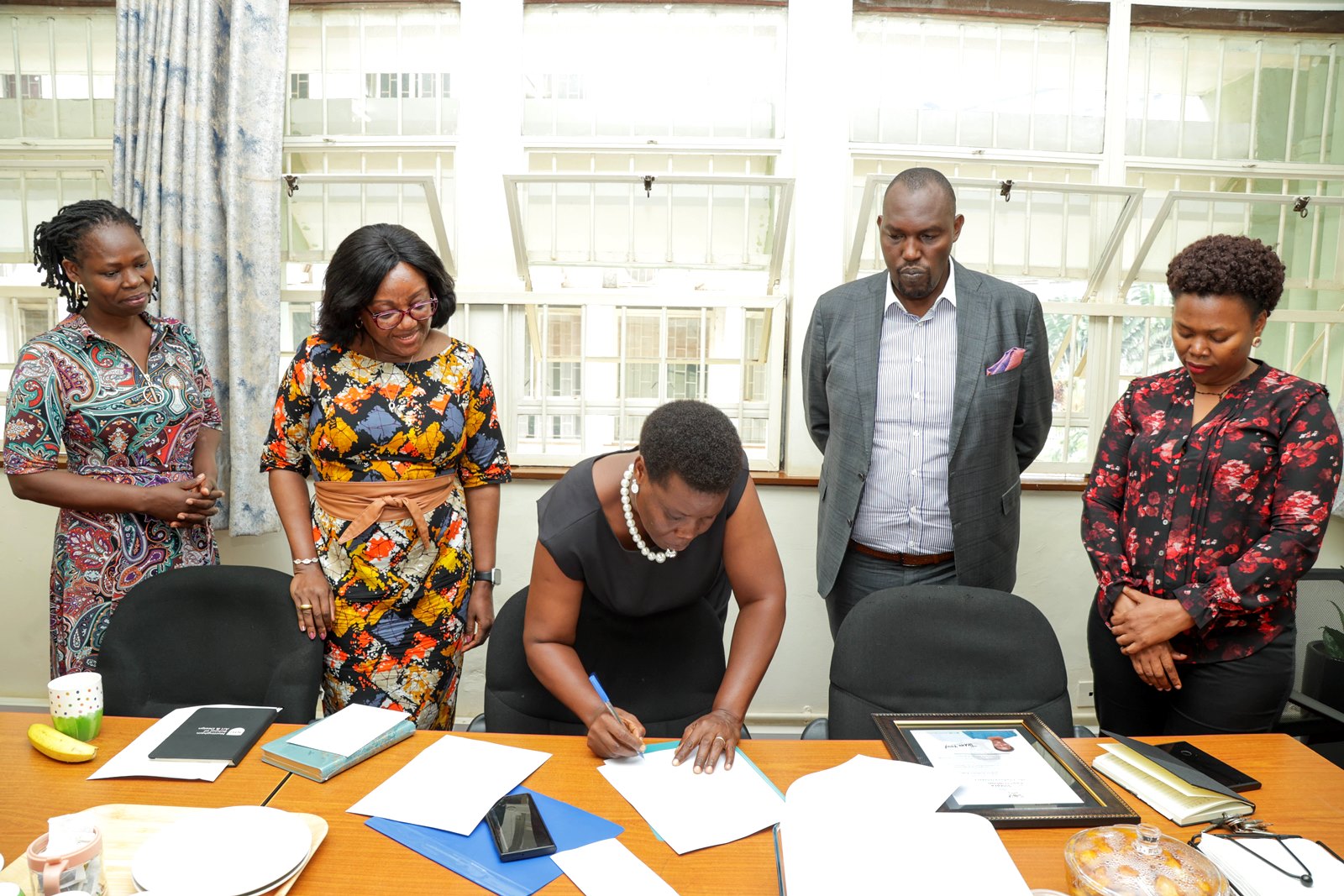 Ms. Gladys Khamili signs her handover report as Dr. Joan Mutyoba, Prof. Rhoda Wanyenze, Mr. Amos Dembe, and incoming Registrar Ms. Annet Khabuya look on. Ms. Gladys Khamili hands over to Ms. Annet Khabuya after 12 years of service as Registrar and transitions to senior role Deputy Registrar, Senate Division, Office of the Academic Registrar during the Makerere University School of Public Health (MakSPH) 239th Management Meeting, 15th April 2026, Mulago Campus, Kampala Uganda, East Africa.