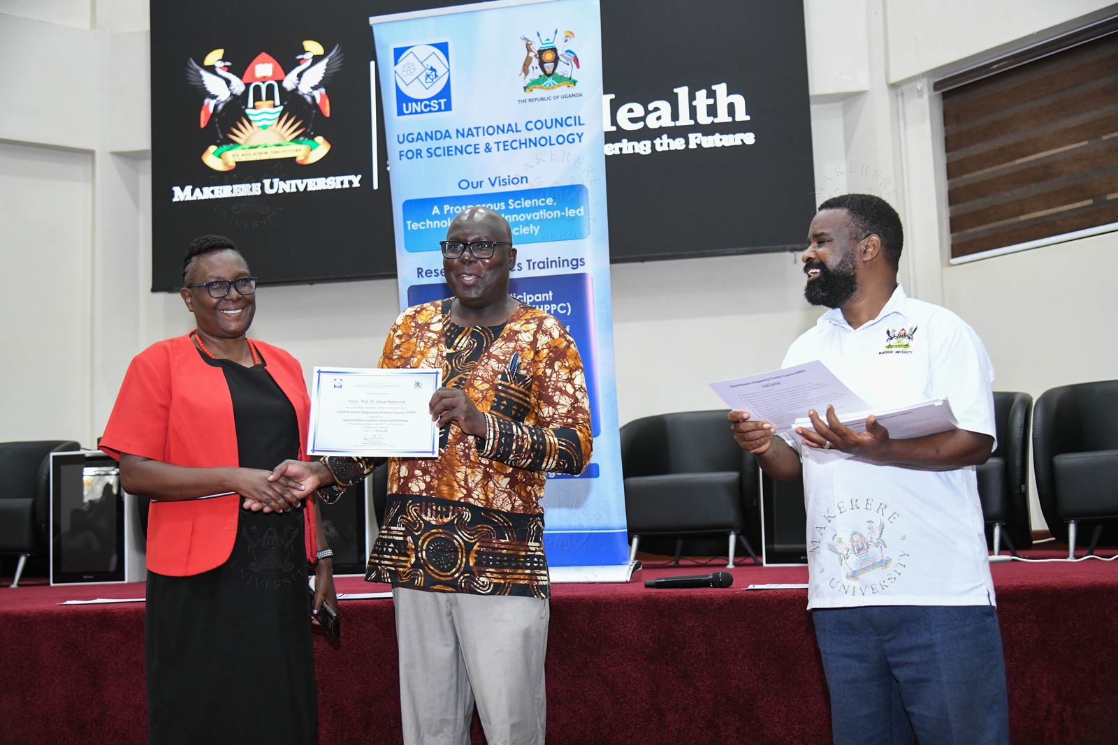 Dr. Martin Patrick Ongol (C) presents a certificate to Prof. Jesca Nakavuma (L) as Prof. Robert Wamala (R) witnesses. Closing Ceremony of the three-day Good Research Regulatory Practice (GRRP) Course for members of Research Ethics Committees (RECs) from Makerere University and other institutions organized by the Directorate of Research, Innovations and Partnerships (DRIP) at Makerere University in partnership with the Uganda National Council for Science and Technology (UNCST), 24th July 2026, MakSPH Auditorium, Kampala Uganda, East Africa.