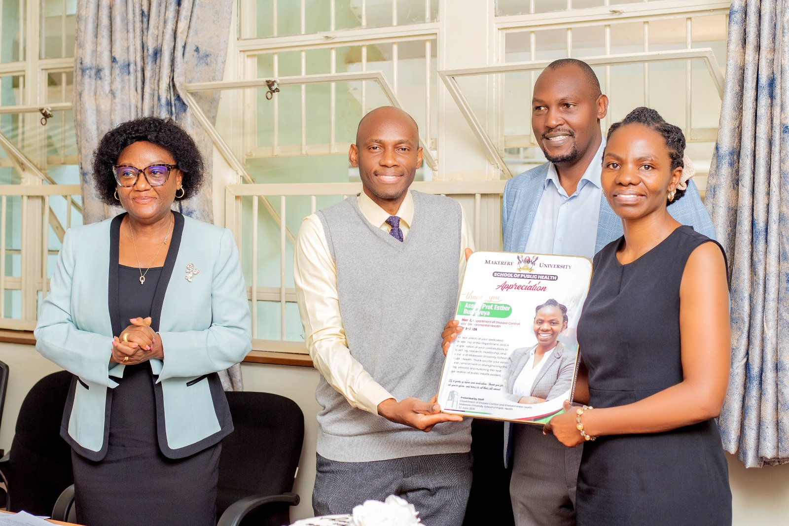 Prof. Rhoda Wanyenze (Left) and Mr. Amos Dembe (Rear) witness as Dr. David Musoke presents an appreciation plaque to Dr. Esther Buregyeya. Makerere University School of Public Health (MakSPH) leadership transition, Department of Disease Control and Environmental Health, Dr. David Musoke takes over from Dr. Esther Buregyeya, 11th August 2026, Mulago Hospital Complex, Kampala Uganda, East Africa.